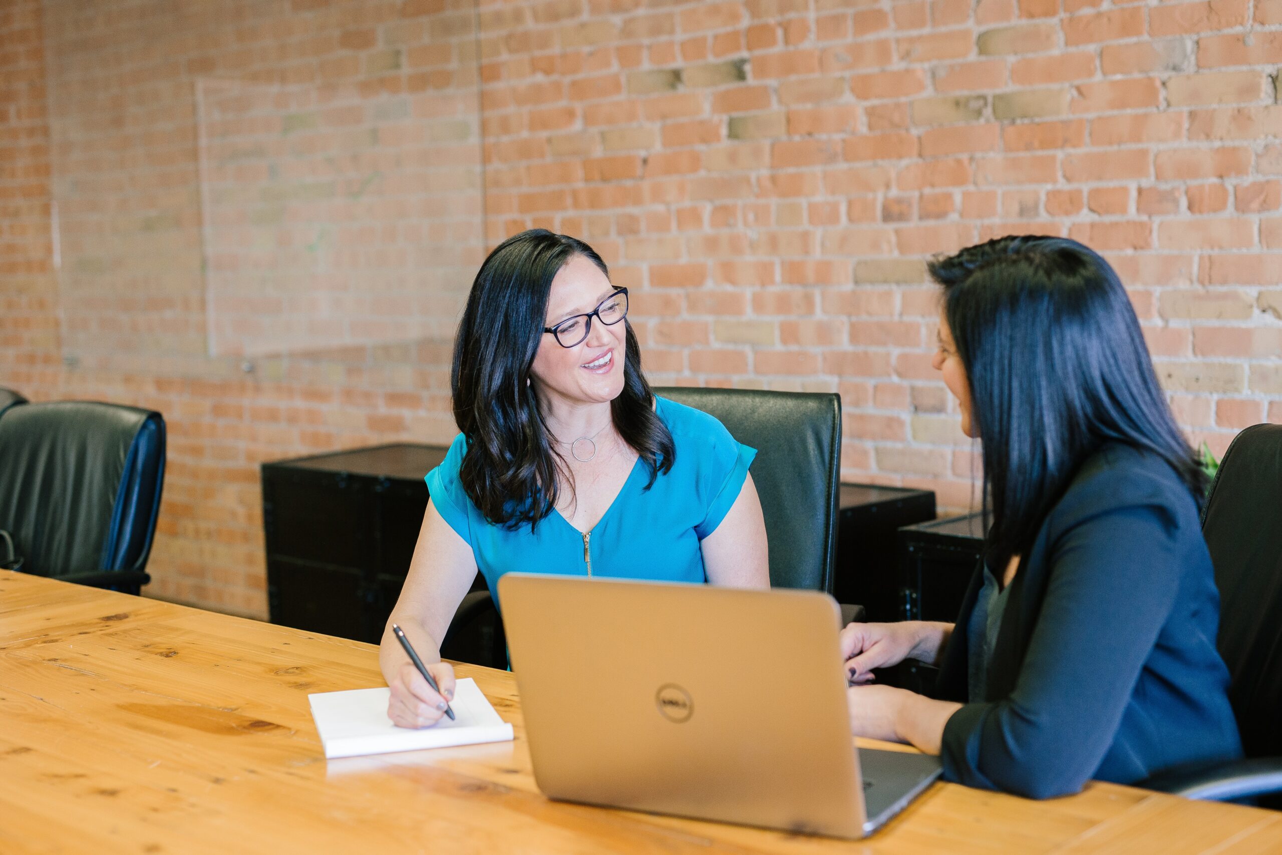 female manager speaking with a female employee at a desk taking notes with an open laptop.